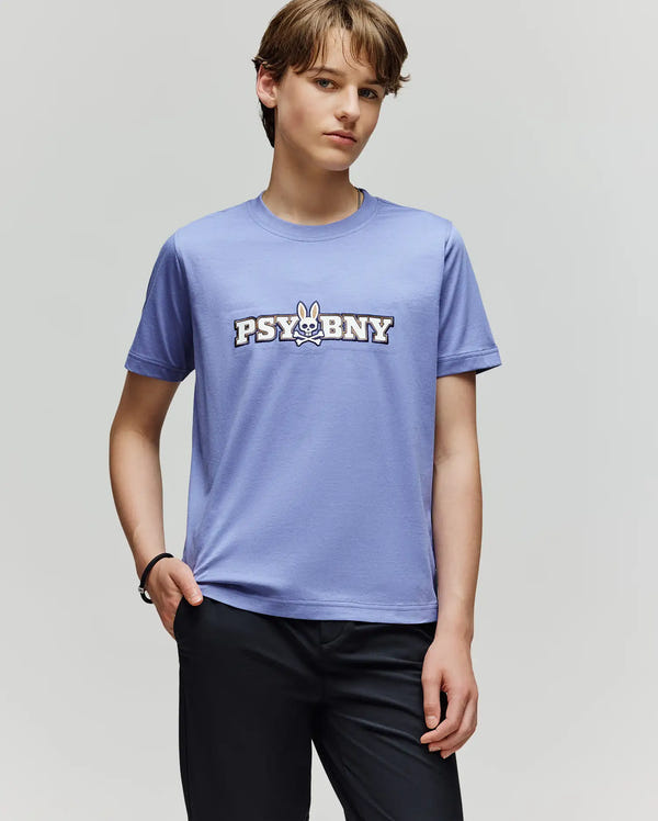 A young person with light brown hair wears the Psycho Bunny KIDS WADE GRAPHIC TEE - KT0200055, a regular fit 100% cotton light purple tee featuring a cartoon bunny and "PSYBNY", paired with dark pants against a plain light gray background.