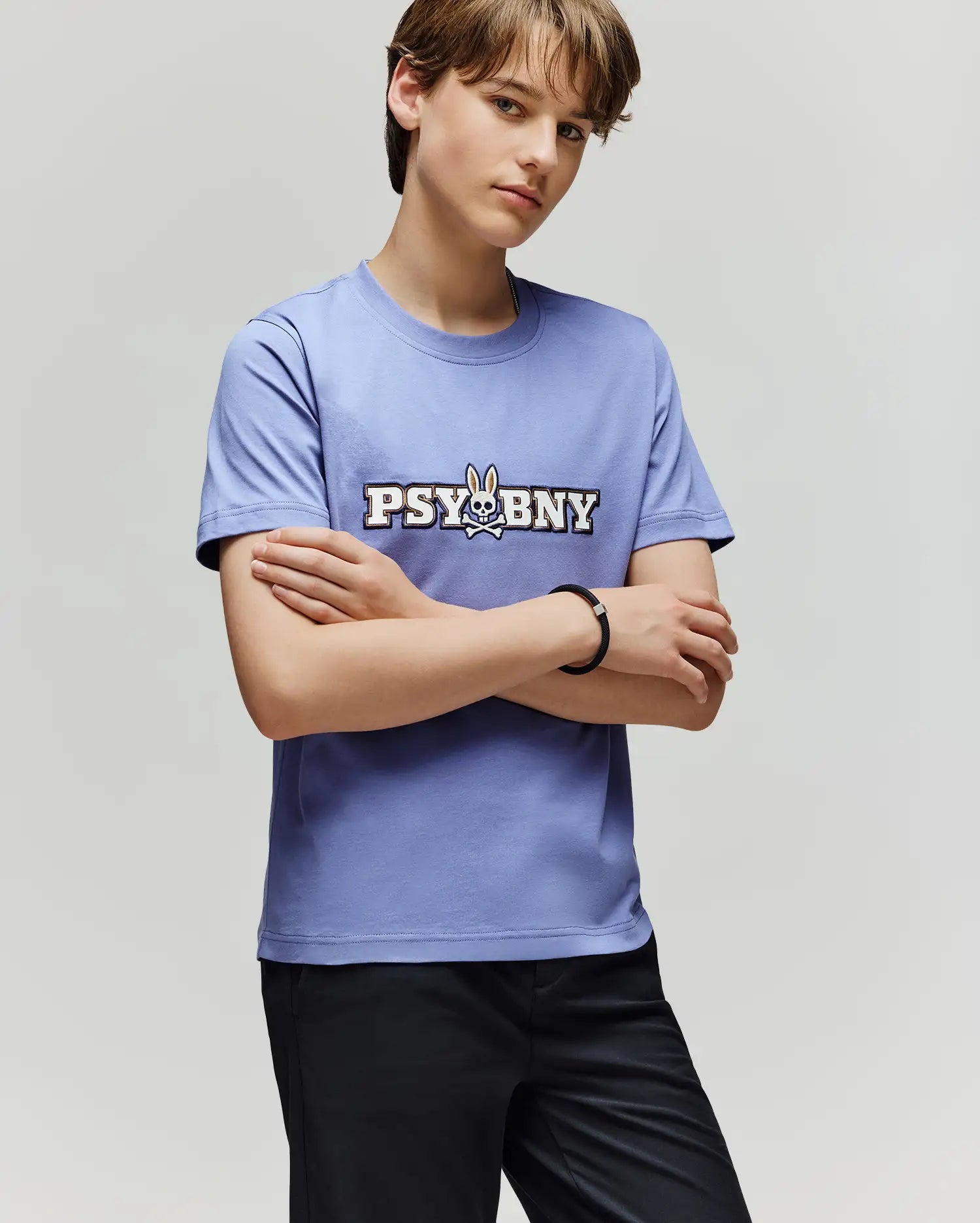 A person with short brown hair stands with arms crossed, wearing the 100% cotton Psycho Bunny KIDS WADE GRAPHIC TEE (KT0200055) in light purple, featuring "PSYBNY" and a bunny graphic, paired with black pants against a plain background.