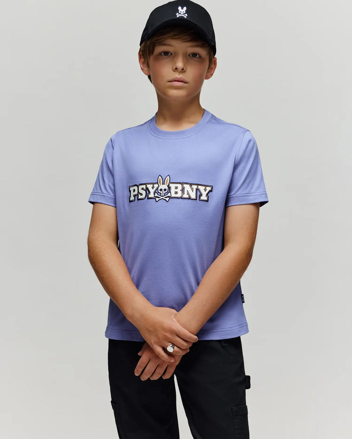 A young boy in a black cap and Psycho Bunny KIDS WADE GRAPHIC TEE (KT0200055) stands against a light gray background, looking at the camera with hands clasped. The lavender tee features 100% cotton, regular fit, cartoon bunny, and "PSYBNY" text.
