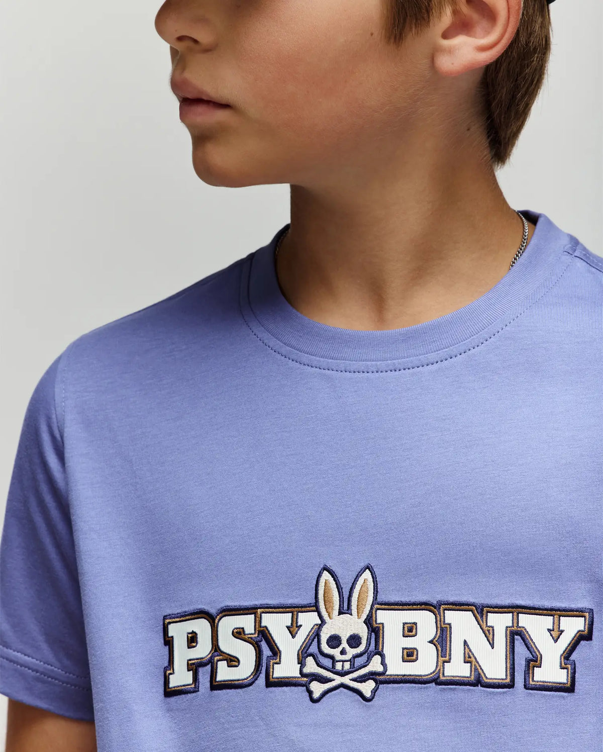 A young person wears the Psycho Bunny KIDS WADE GRAPHIC TEE (KT0200055) in light purple, featuring "PSYBUNY" and a bunny-eared skull-and-crossbones logo on the chest. Part of their face is partially out of frame.