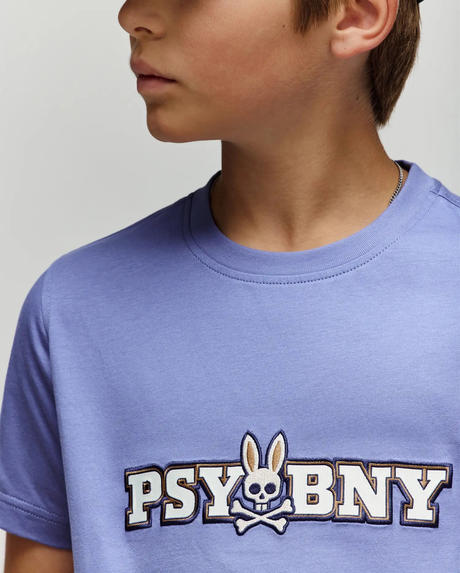A young person wears the Psycho Bunny KIDS WADE GRAPHIC TEE (KT0200055) in light purple, featuring "PSYBUNY" and a bunny-eared skull-and-crossbones logo on the chest. Part of their face is partially out of frame.