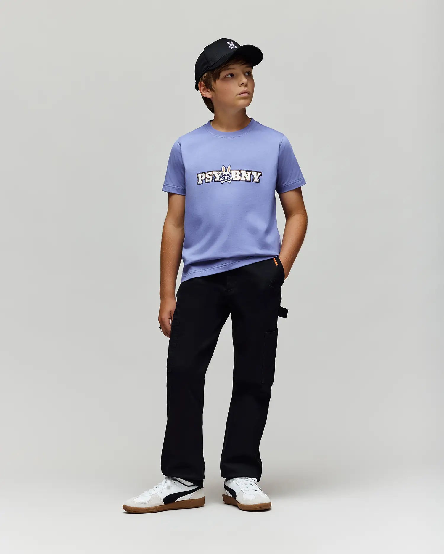 A boy in a black baseball cap, Psycho Bunny KIDS WADE GRAPHIC TEE - KT0200055 (light purple with "PSYBNY" and cartoon bunny), regular fit black pants, and white sneakers stands against a light gray background.