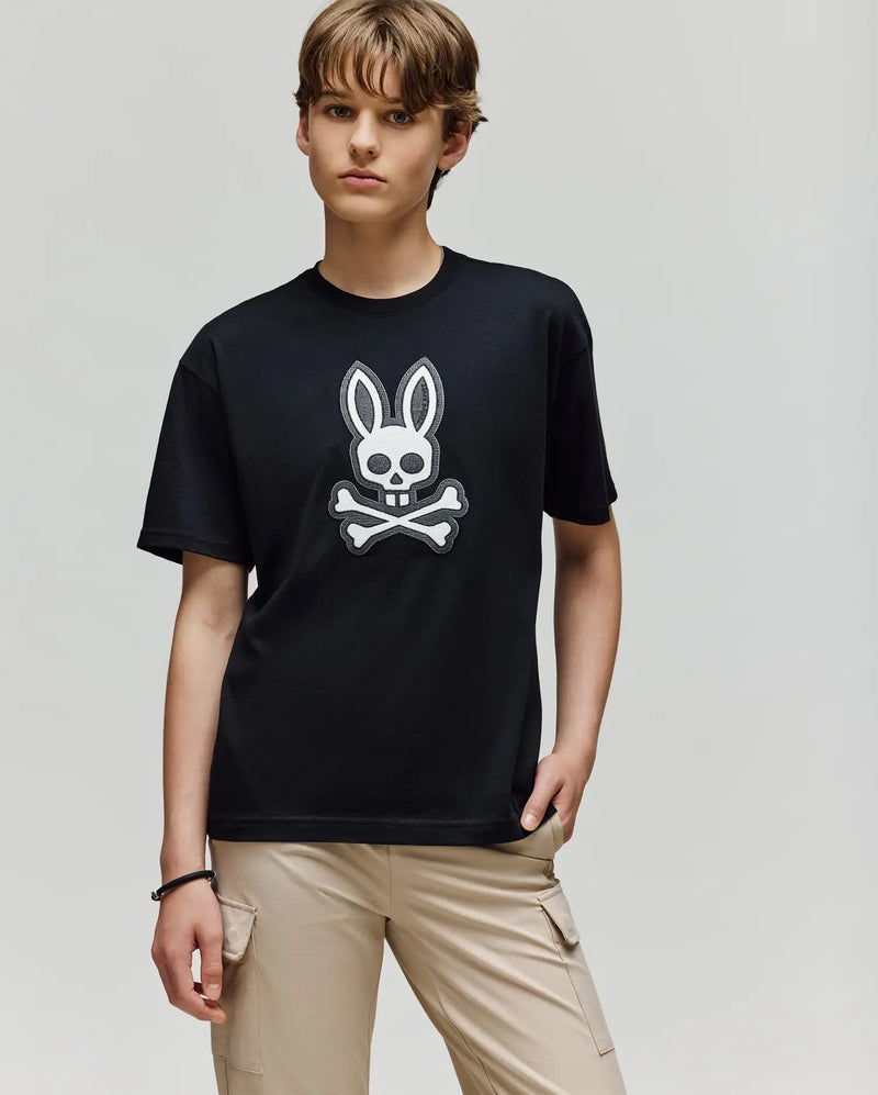 A young person wears the Psycho Bunny KIDS RYDER OVERSIZED FIT TEE (KT0200056), featuring an embroidered Bunny skull and crossbones graphic, with one hand in their beige cargo pants pocket, standing against a plain light background.