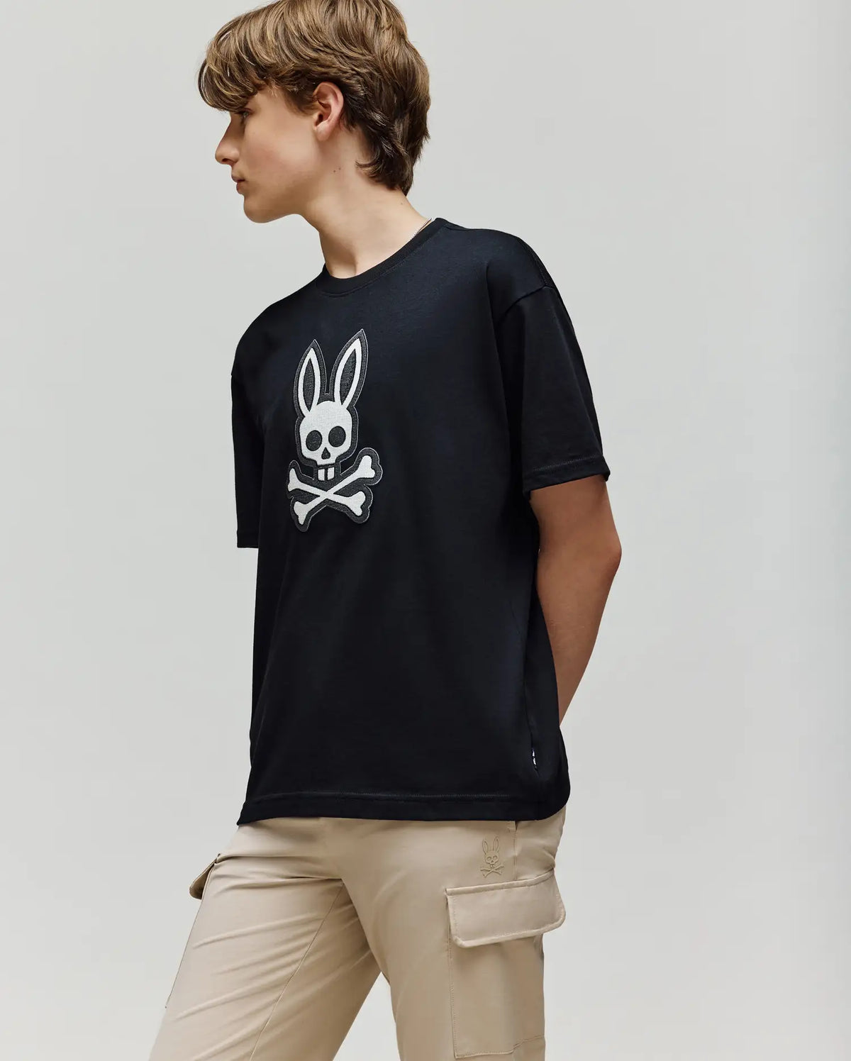 A person with short brown hair wears the Psycho Bunny KIDS RYDER OVERSIZED FIT TEE (KT0200056), featuring an embroidered Bunny skull and crossbones logo, paired with beige cargo pants, standing against a plain light background.