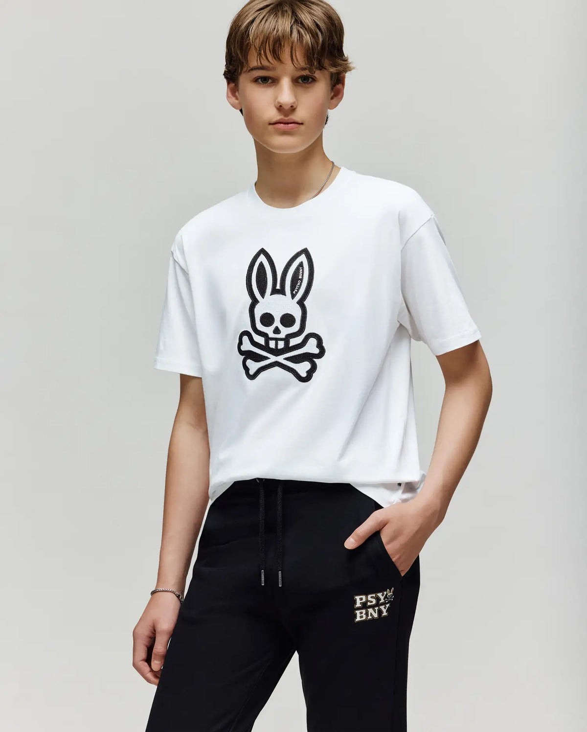 A person with short brown hair wears the Psycho Bunny KIDS RYDER OVERSIZED FIT TEE (KT0200056) with a black embroidered Bunny skull graphic, paired with black "PSY BNXY" pants, standing against a plain light background.