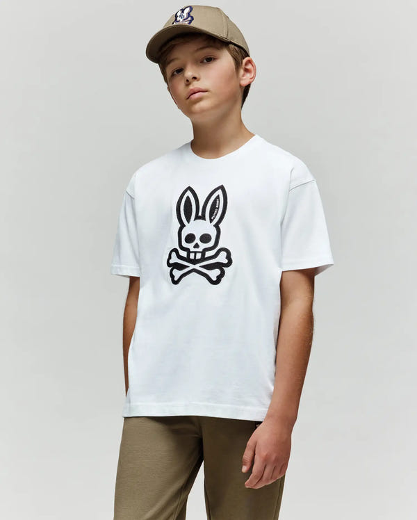 A boy in khaki pants and a beige cap wears the Psycho Bunny KIDS RYDER OVERSIZED FIT TEE (KT0200056), featuring a large black bunny skull and crossbones graphic, standing against a plain light gray background.