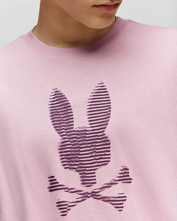 Wearing the Psycho Bunny KIDS GRANT BUNNY GRAPHIC TEE KT0200061, a person shows only their lower face and upper torso in this HD-printed light pink Pima cotton tee featuring a dark purple striped bunny skull graphic.
