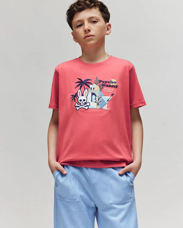 A boy stands with hands in the pockets of his blue shorts, wearing the Psycho Bunny KIDS SHANE GRAPHIC TEE (KT0200090), a coral pink Pima cotton shirt with a cartoon bunny, palm tree, cactus, and "Psycho Bunny" text on the front. The background is light gray.