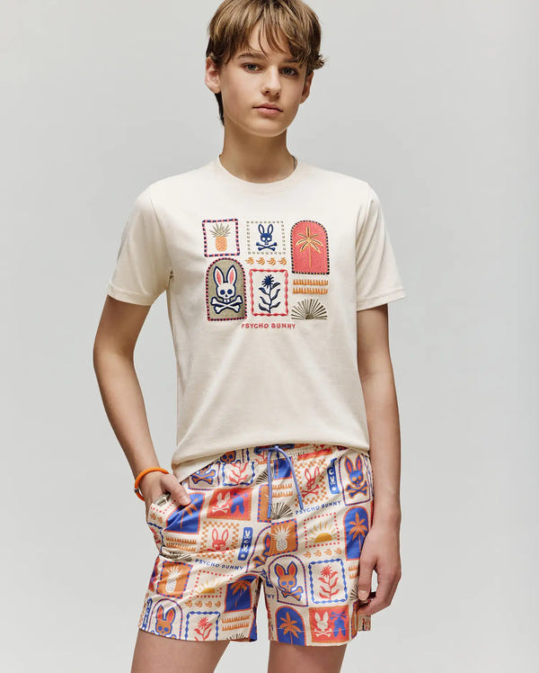 A teen wears the Psycho Bunny KIDS SUNDEW GRAPHIC TEE (KT0200092), a cream Pima cotton top with colorful squares of bunnies and plants, paired with matching vibrant shorts for a fresh ocean view vibe.