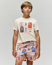 A young boy stands against a plain background wearing the Psycho Bunny KIDS SUNDEW GRAPHIC TEE (KT0200092), a cream Pima cotton shirt with colorful graphics, paired with shorts featuring red, blue, and orange Beach Bunny print motifs.