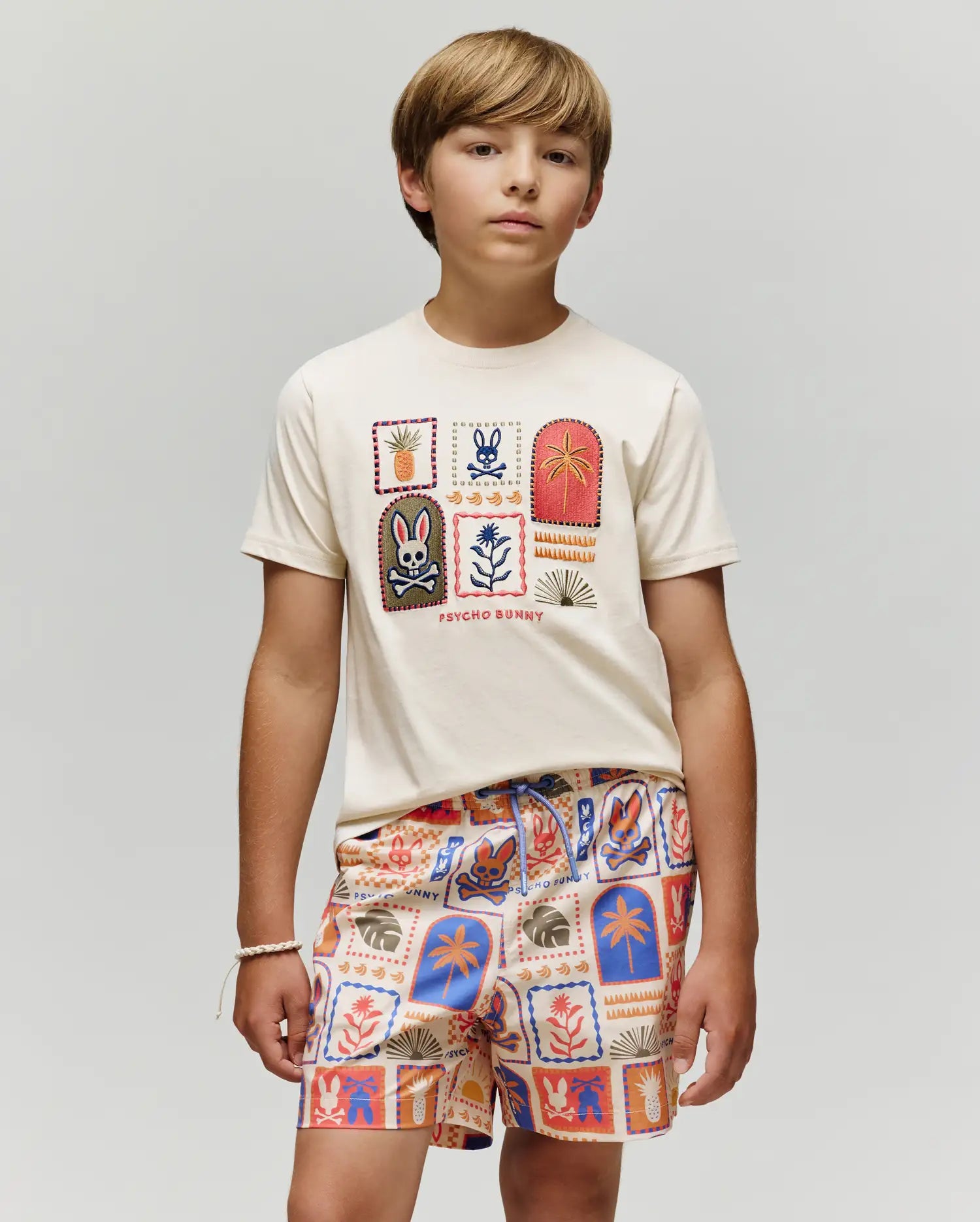 A young boy stands against a plain background wearing the Psycho Bunny KIDS SUNDEW GRAPHIC TEE (KT0200092), a cream Pima cotton shirt with colorful graphics, paired with shorts featuring red, blue, and orange Beach Bunny print motifs.