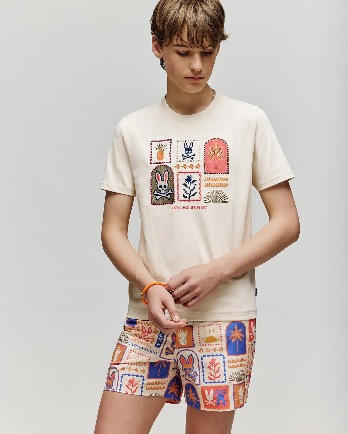 A child wears the Psycho Bunny KIDS SUNDEW GRAPHIC TEE (KT0200092), a cream Pima cotton shirt with colorful Beach Bunny prints, paired with matching playful shorts, standing against a plain light background and looking down.