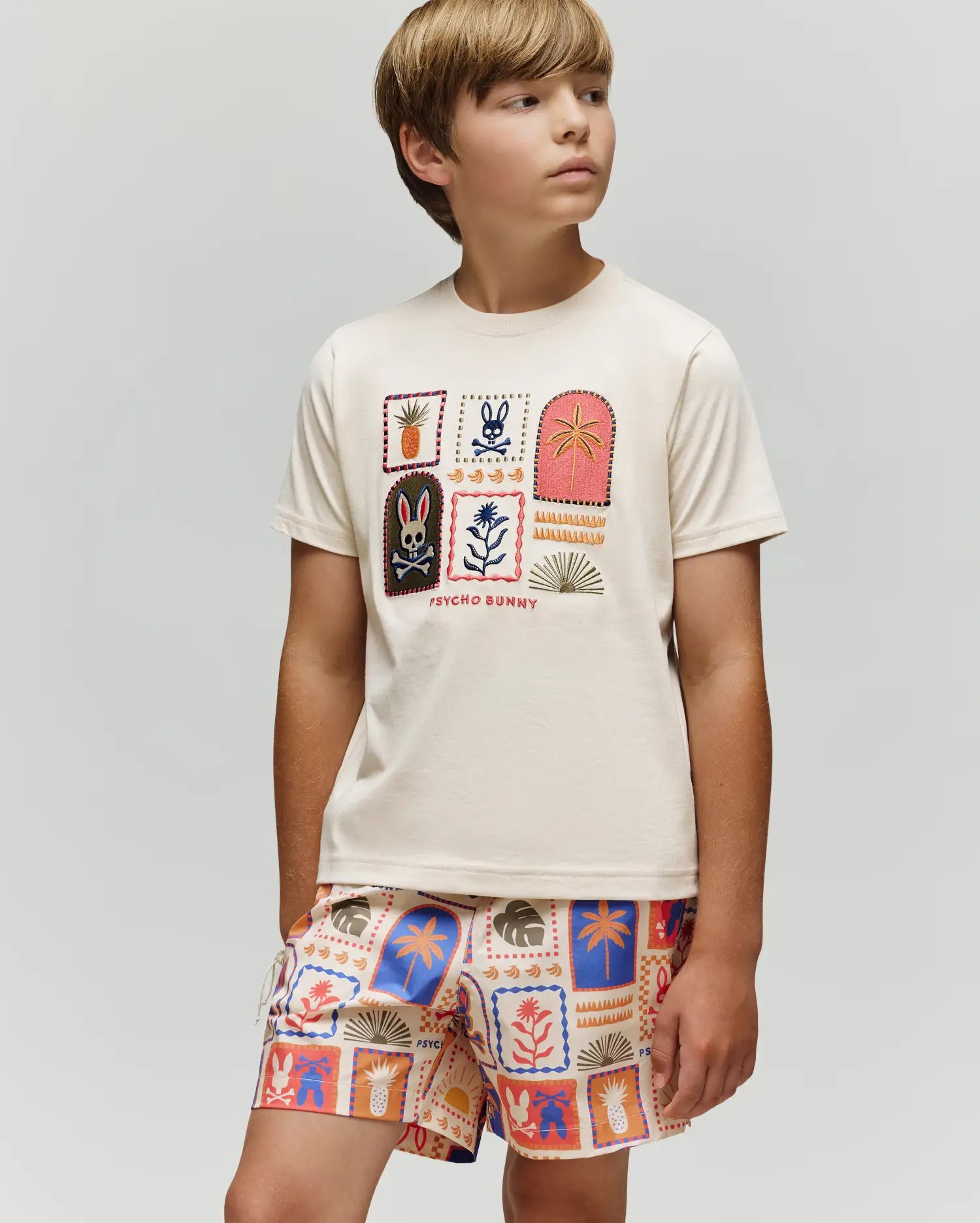 A boy in the Psycho Bunny KIDS SUNDEW GRAPHIC TEE (KT0200092), a cream Pima cotton shirt with colorful Beach Bunny prints, paired with matching shorts, stands against a plain light background, gazing slightly sideways.