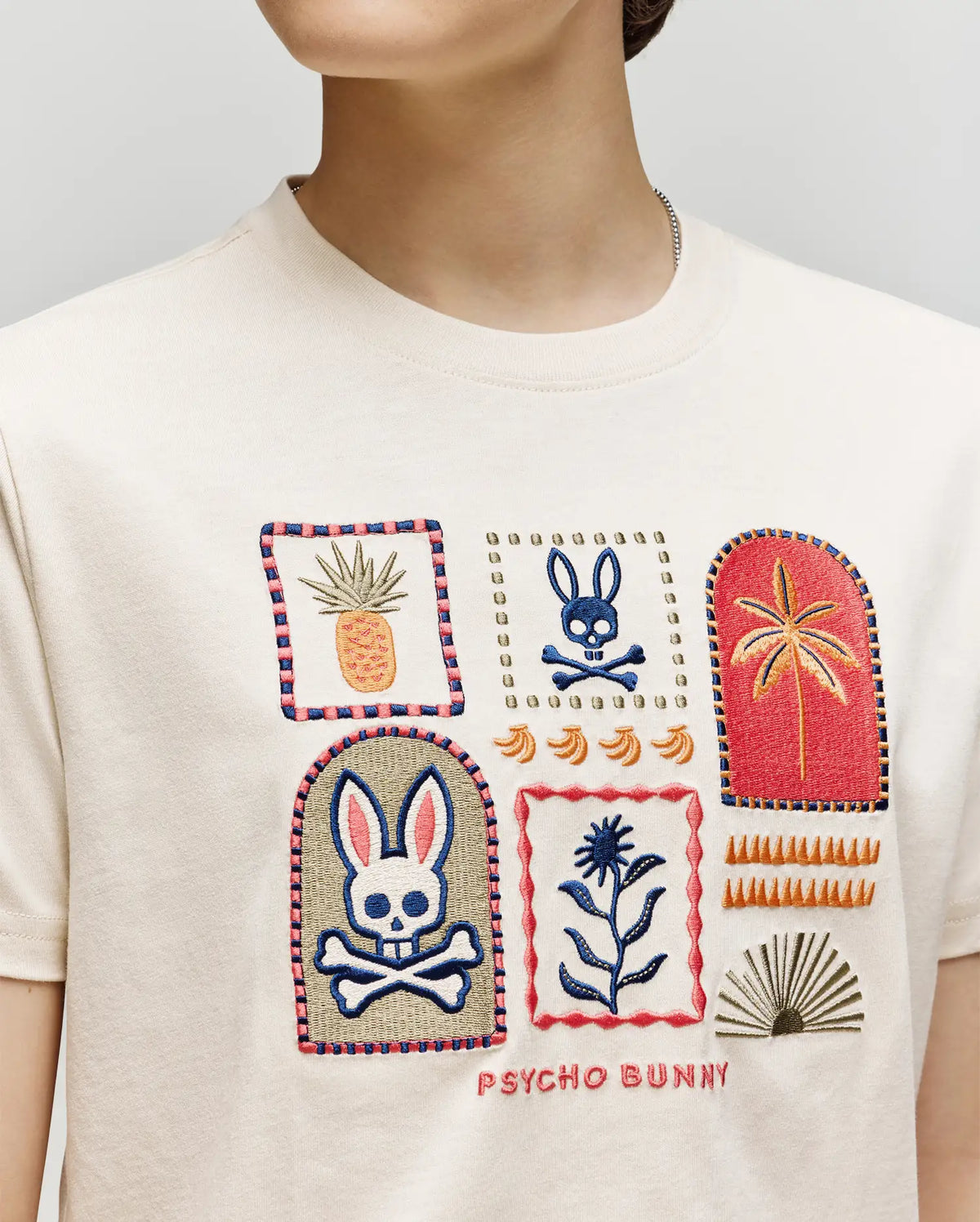 A child wears the Psycho Bunny KIDS SUNDEW GRAPHIC TEE (KT0200092), a cream Pima cotton shirt with colorful embroidered bunnies, skulls, plants, geometric shapes, and "PSYCHO BUNNY" stitched at the hem for an ocean-inspired flair.