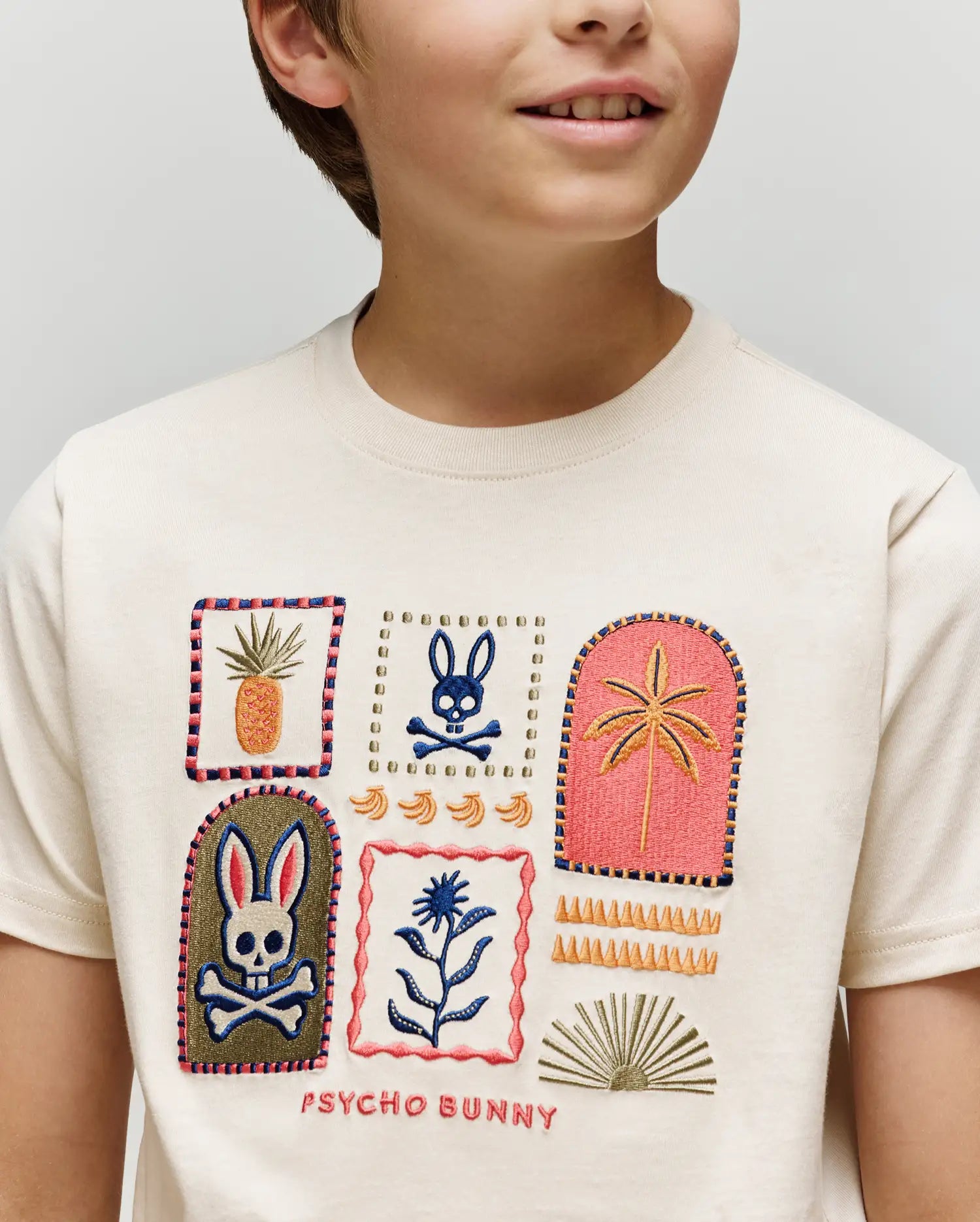 A child wears the Psycho Bunny KIDS SUNDEW GRAPHIC TEE (KT0200092), a light Pima cotton shirt featuring embroidered bunnies, skulls, pineapple, plants, decorative shapes, and "Psycho Bunny" text for an ocean-inspired style.