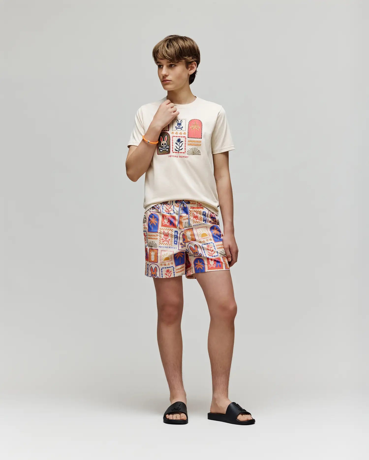 A young person stands against a light background, wearing the Psycho Bunny KIDS SUNDEW GRAPHIC TEE (KT0200092) with colorful Beach Bunny designs, patterned shorts, and black slide sandals, looking to the side with one hand near their chin.