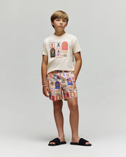 A boy stands against a light gray background, wearing the Psycho Bunny KIDS SUNDEW GRAPHIC TEE (KT0200092), patterned ocean-inspired shorts, black slide sandals, and a white bracelet on his wrist.