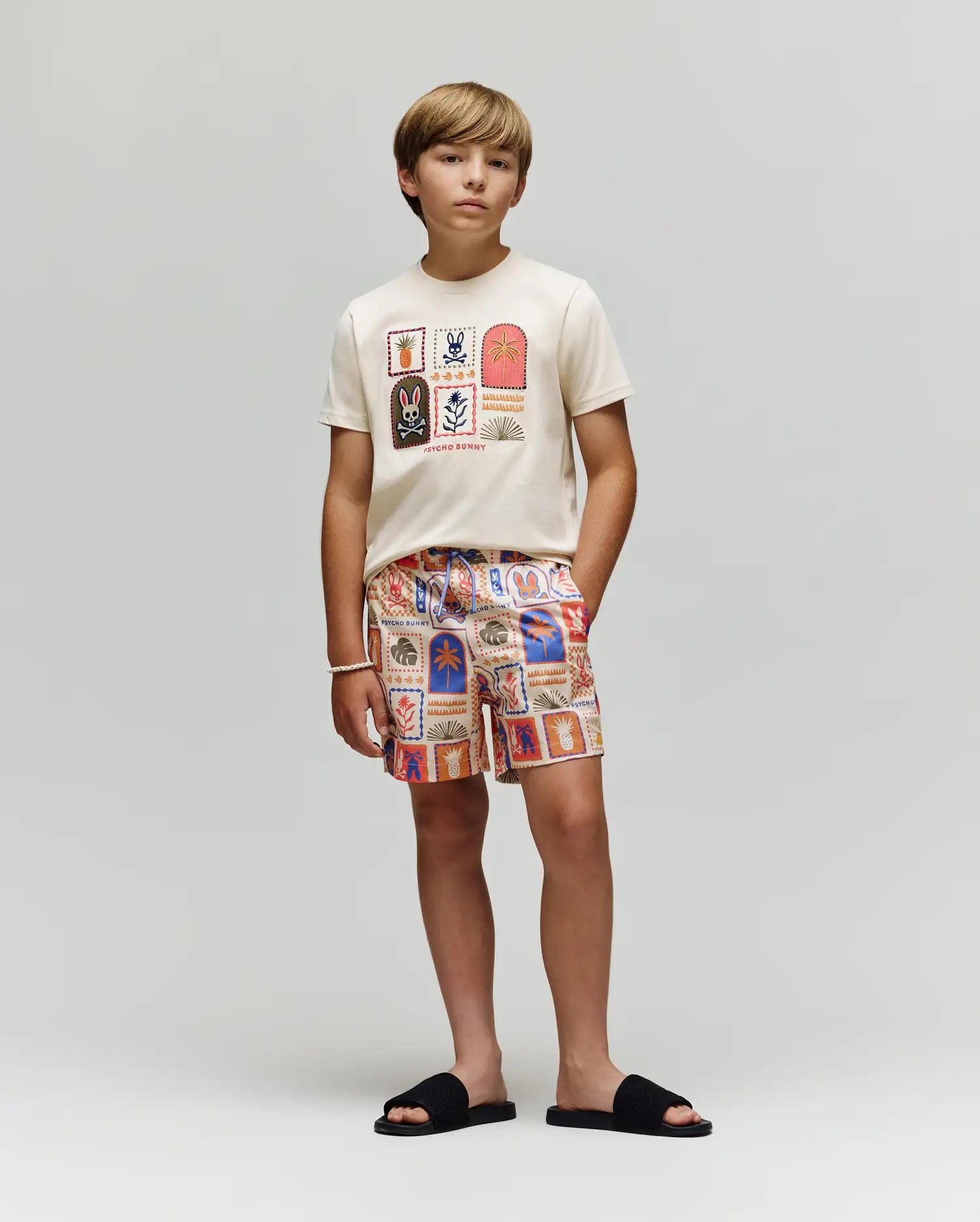 A boy stands against a light gray background, wearing the Psycho Bunny KIDS SUNDEW GRAPHIC TEE (KT0200092), patterned ocean-inspired shorts, black slide sandals, and a white bracelet on his wrist.