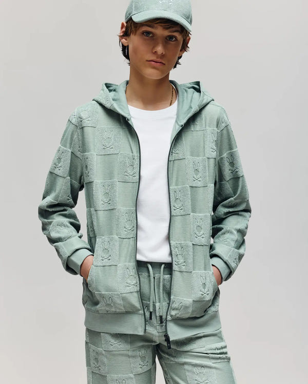 A young person wears the Psycho Bunny KIDS KELVIN FULL ZIP TOWEL TERRY HOODIE (KT0500004) in light green, with matching joggers and a cap, hands in pockets, standing against a plain light background.