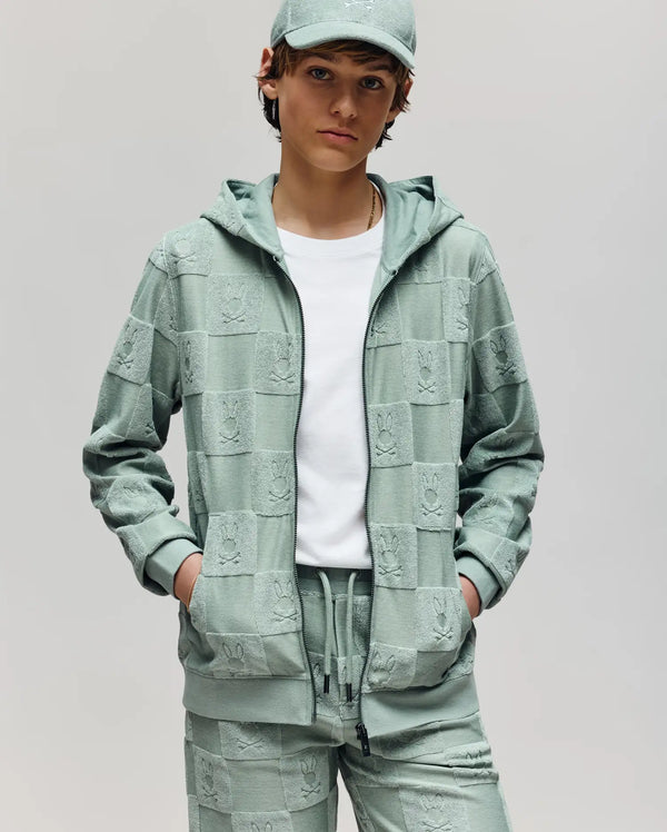 A young person wears the Psycho Bunny KIDS KELVIN FULL ZIP TOWEL TERRY HOODIE (KT0500004) in light green, with matching joggers and a cap, hands in pockets, standing against a plain light background.