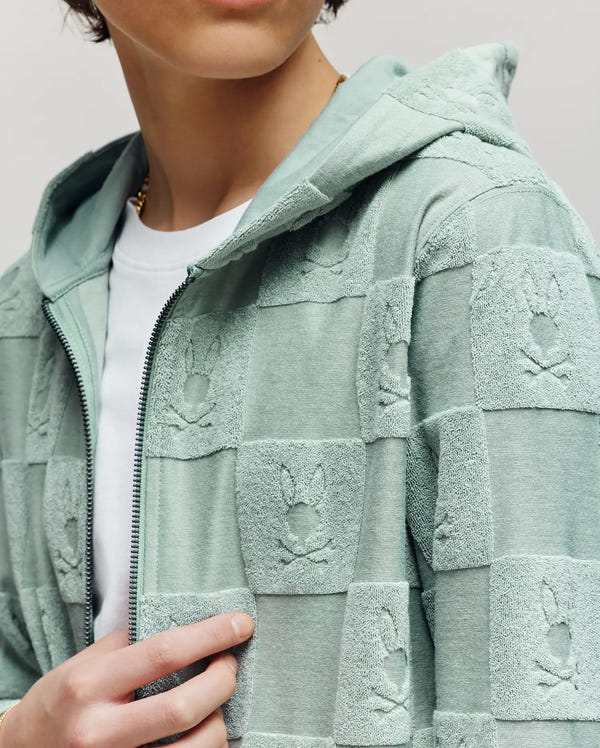 A person wears the Psycho Bunny KIDS KELVIN FULL ZIP TOWEL TERRY HOODIE (KT0500004), a mint green cotton-blend jacquard terry zip-up with a checkered bunny pattern and hood, layered over a white t-shirt. Face is partially out of frame.