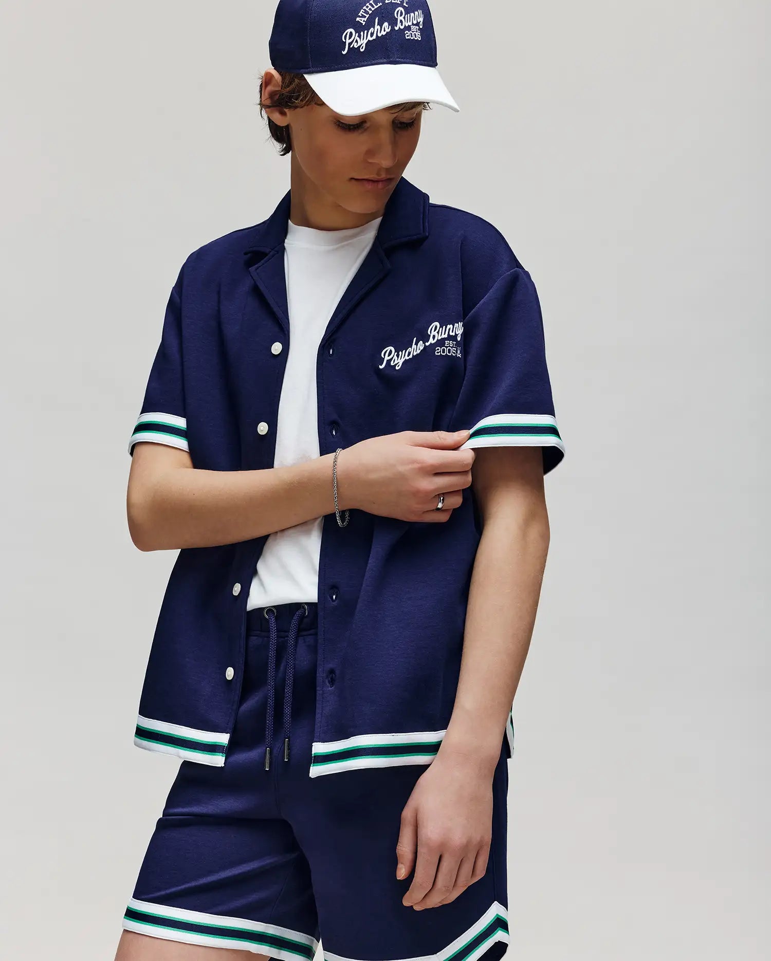 A child wears the Psycho Bunny KIDS BASIL TAPED CAMP COLLAR SHIRT (KT0500007), a navy blue cotton-modal blend shirt with white and green striped trim, "Psycho Bunny" on the chest, paired with a white tee and navy cap while adjusting their sleeve.