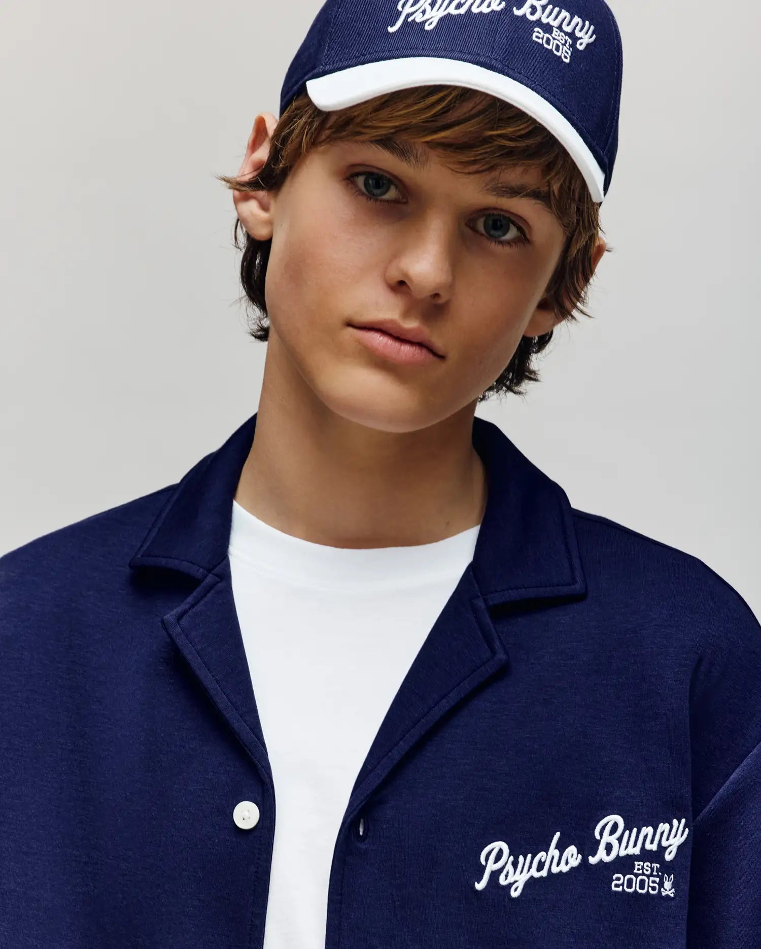 A young person in a navy Psycho Bunny cap and jacket wears the KIDS BASIL TAPED CAMP COLLAR SHIRT - KT0500007, a premium cotton-modal blend white shirt by Psycho Bunny, looking neutrally at the camera against a light plain background.