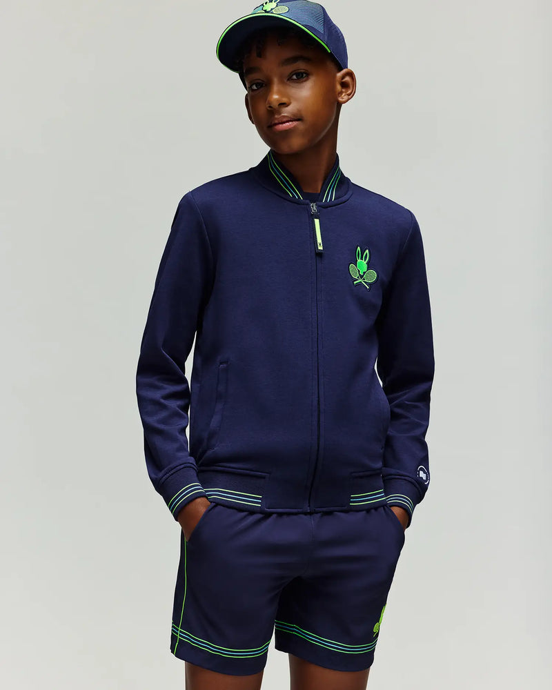 A young boy wears the Psycho Bunny RETAIL BALL CREW YOUTH JACKET (EN) - KT0500014 in navy blue with green accents and matching shorts, plus a navy blue cap, standing with hands in his pockets against a plain light background.