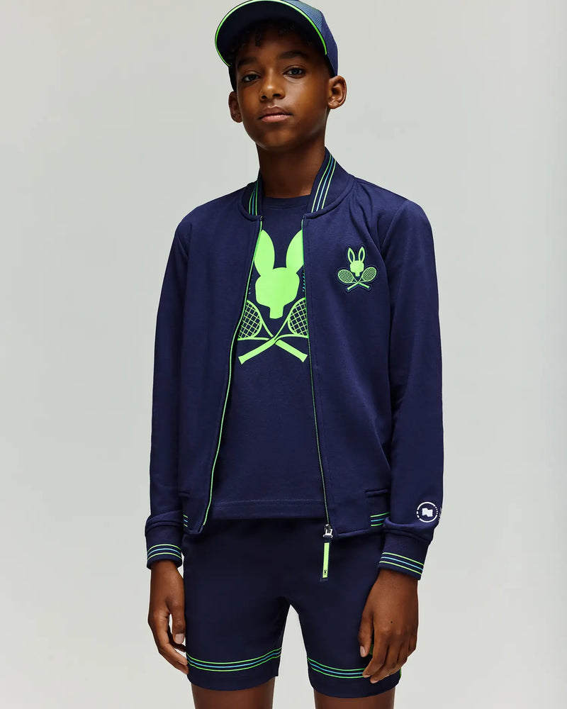 A boy stands facing forward against a plain background, wearing the Psycho Bunny RETAIL BALL CREW YOUTH JACKET (EN) - KT0500014, a navy blue cap, a neon green tennis graphic t-shirt, and matching shorts.