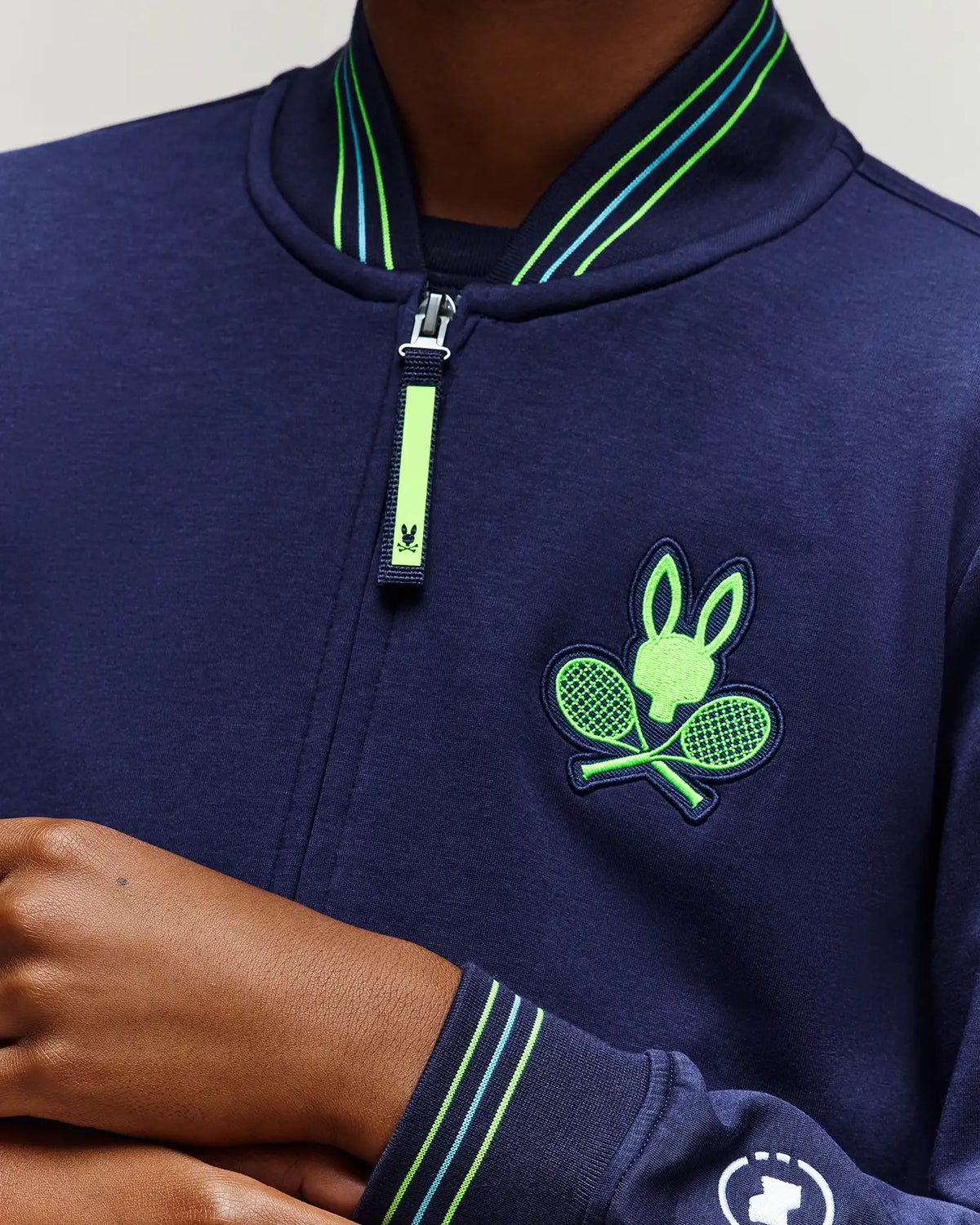 A person is wearing the Psycho Bunny RETAIL BALL CREW YOUTH JACKET (EN) - KT0500014: a navy jacket with green striped trim, a zipper, and a chest logo showing a rabbit head and crossed tennis rackets. Only their lower face, neck, and hand are visible.