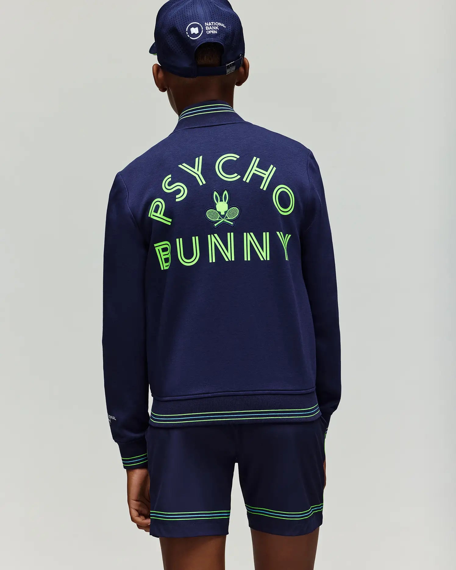 A person stands facing away, wearing the Psycho Bunny RETAIL BALL CREW YOUTH JACKET (EN) - KT0500014 in navy with bold neon "PSYCHO BUNNY" lettering and bunny logo, paired with navy shorts and a matching cap for a sporty, casual look.