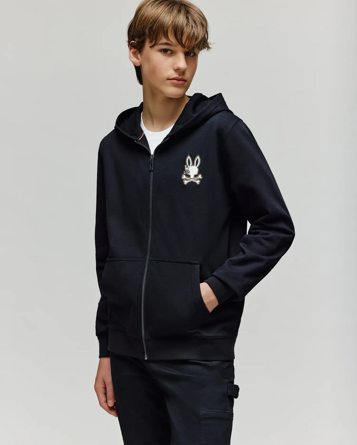 A young person stands with one hand in their pocket against a light background, wearing the Psycho Bunny KIDS WADE CORD PATCH ZIP HOODIE (KT0500016) in black, featuring a small bunny and crossbones chest design.