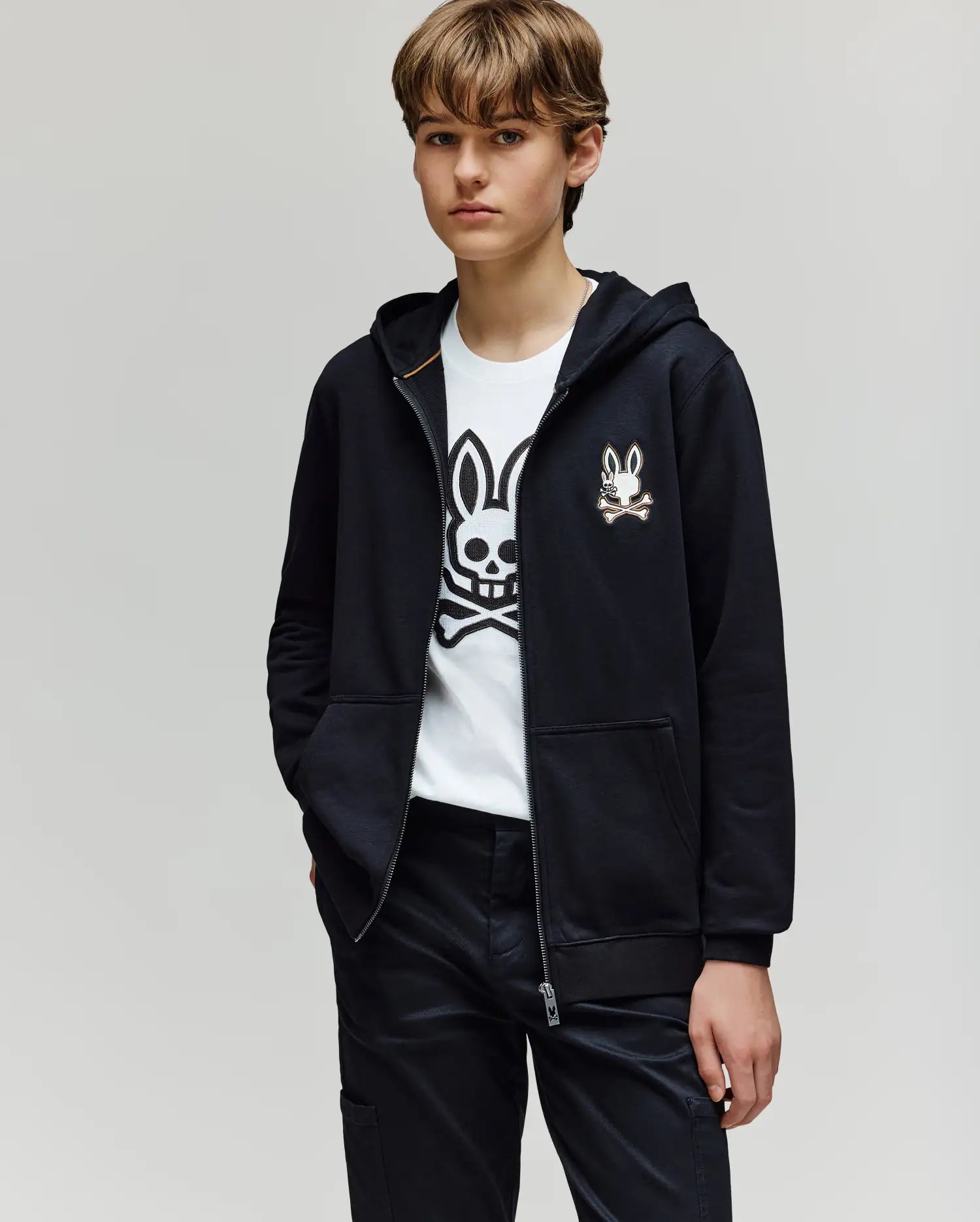 A young person with short brown hair wears the Psycho Bunny KIDS WADE CORD PATCH ZIP HOODIE (KT0500016) over a white T-shirt with a cartoon bunny skull logo, standing with one hand in their pocket against a plain light background.