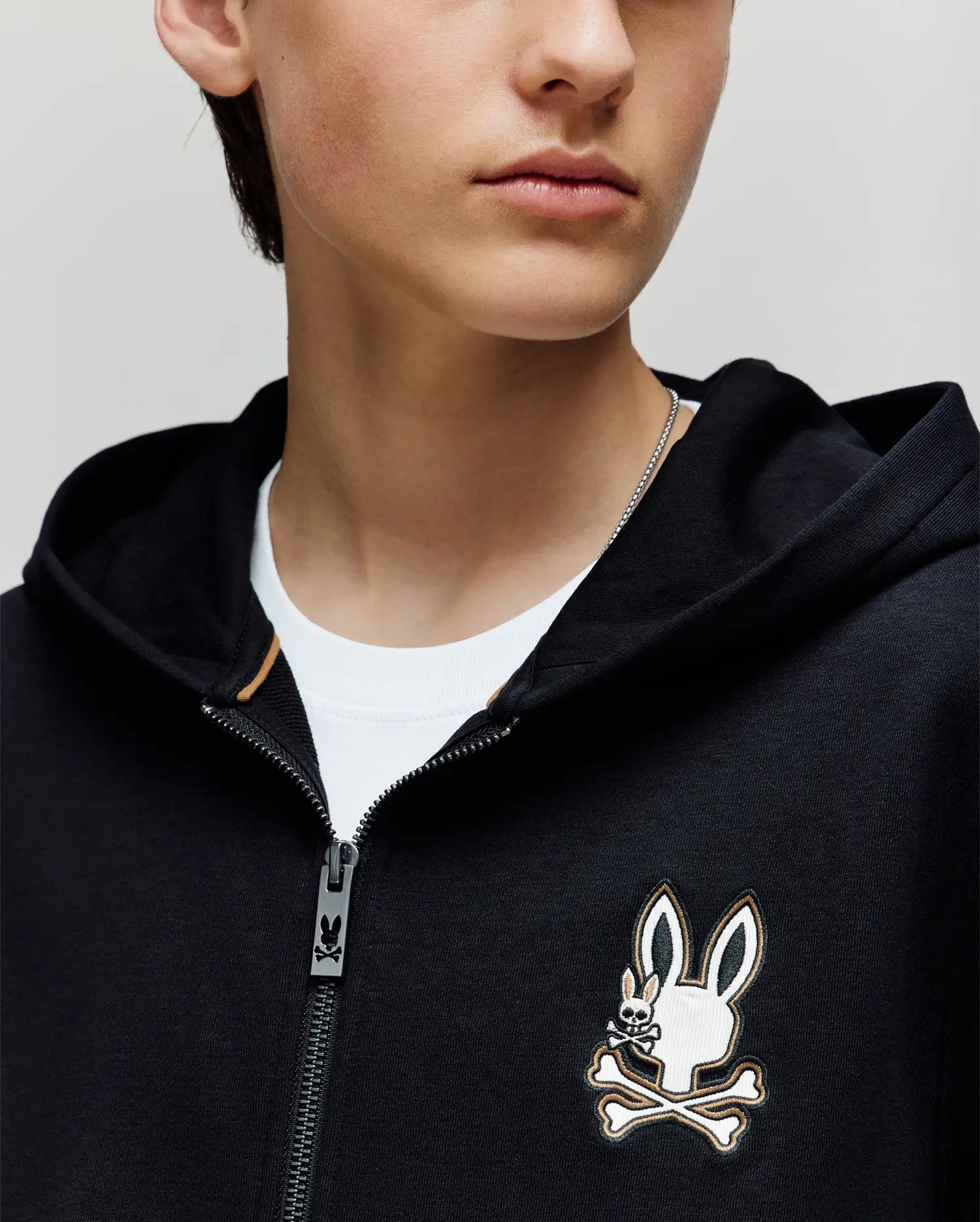 A person in a Psycho Bunny KIDS WADE CORD PATCH ZIP HOODIE (KT0500016)—a black premium cotton zip-hoodie with a cartoon bunny and crossbones chest logo—layered over a white t-shirt. Photo shows only the lower face and upper torso.