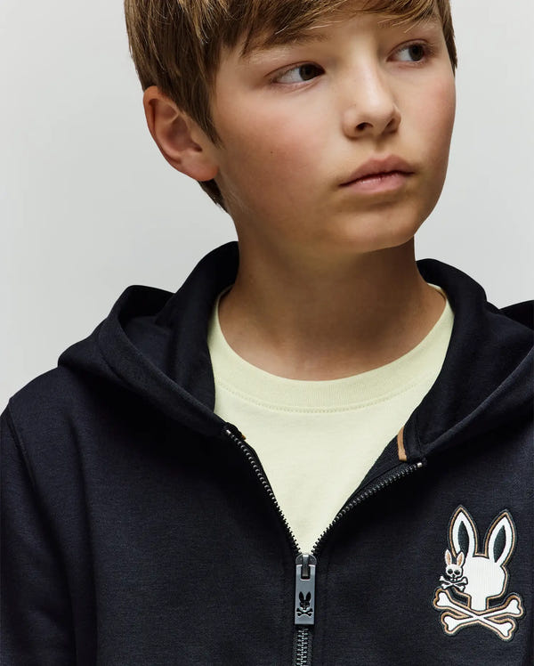 A young boy with light brown hair looks to the side, wearing the Psycho Bunny KIDS WADE CORD PATCH ZIP HOODIE (KT0500016) in black, featuring a cartoon bunny and crossbones logo, premium cotton blend, and an embroidered hood over a cream T-shirt.