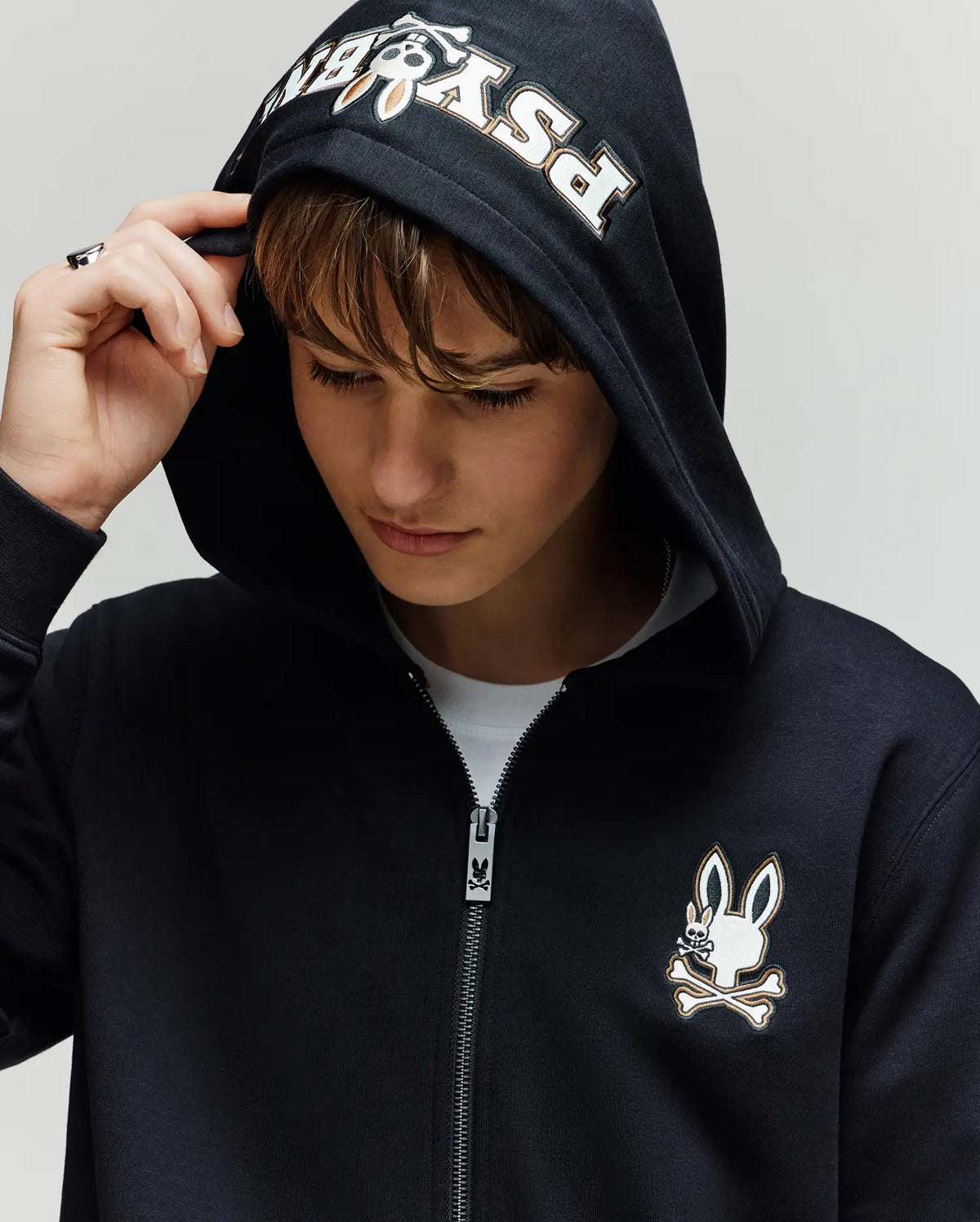 A person wears the Psycho Bunny KIDS WADE CORD PATCH ZIP HOODIE (KT0500016), a black cotton blend zip hoodie with a stylized bunny and crossbones logo on the chest and "Psycho" embroidered on the hood, looking down neutrally.