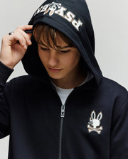 A person wears the Psycho Bunny KIDS WADE CORD PATCH ZIP HOODIE (KT0500016), a black cotton blend zip hoodie with a stylized bunny and crossbones logo on the chest and "Psycho" embroidered on the hood, looking down neutrally.