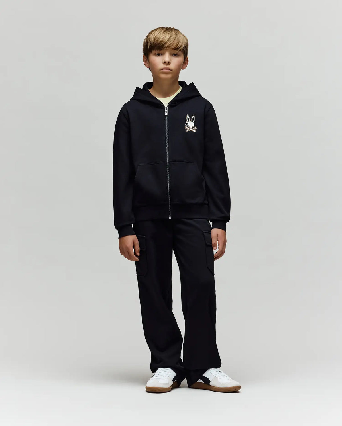 A young boy faces forward in a Psycho Bunny KIDS WADE CORD PATCH ZIP HOODIE (KT0500016) made from a premium cotton blend with a small cartoon graphic on the chest, paired with black cargo pants and white sneakers against a light gray background.