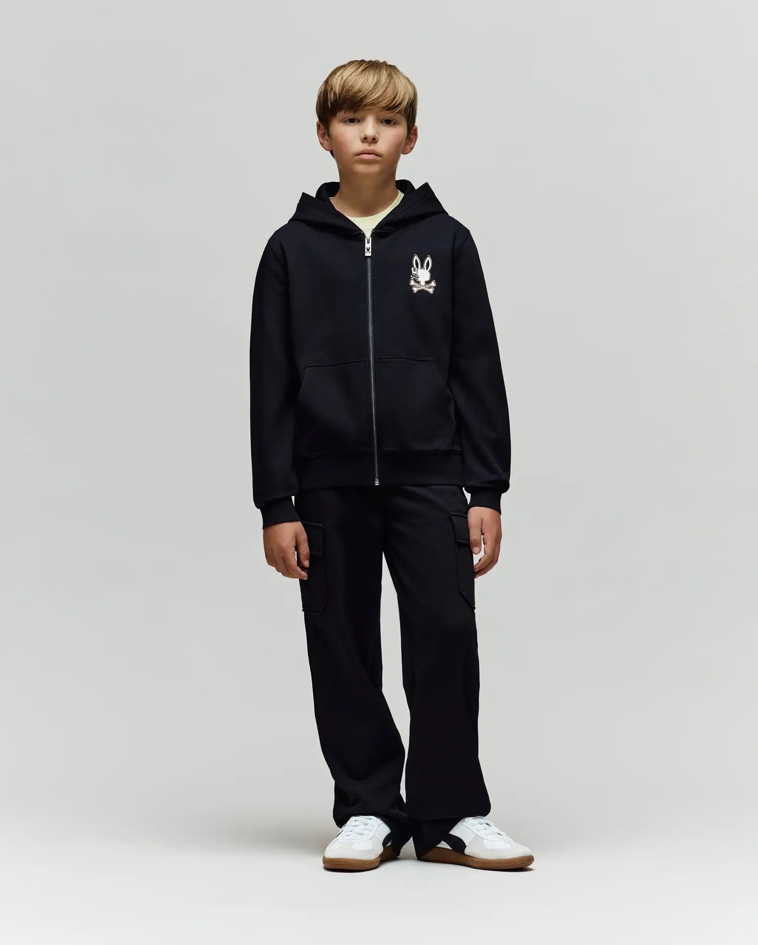 A young boy faces forward in a Psycho Bunny KIDS WADE CORD PATCH ZIP HOODIE (KT0500016) made from a premium cotton blend with a small cartoon graphic on the chest, paired with black cargo pants and white sneakers against a light gray background.