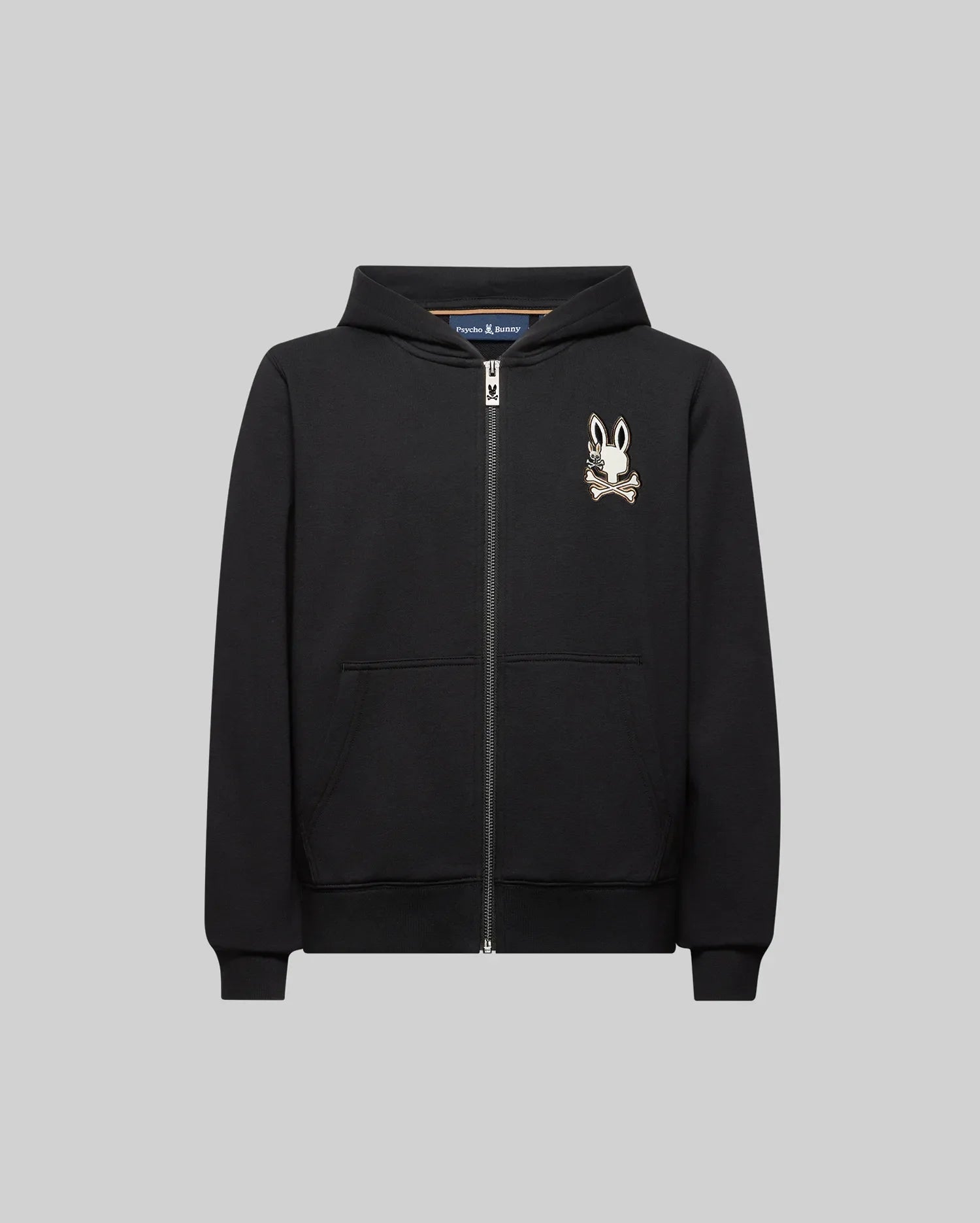 The Psycho Bunny KIDS WADE CORD PATCH ZIP HOODIE (KT0500016) is a black zip-up hoodie crafted from a premium cotton blend, featuring a hood, front pockets, and an embroidered bunny skull logo on the left chest. Displayed on a plain grey background.