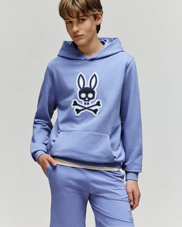 A young person wears the Psycho Bunny KIDS RYDER EMBROIDERED BUNNY PATCH HOODIE (KT0500018) in light blue, paired with matching loungewear shorts, against a plain light background.