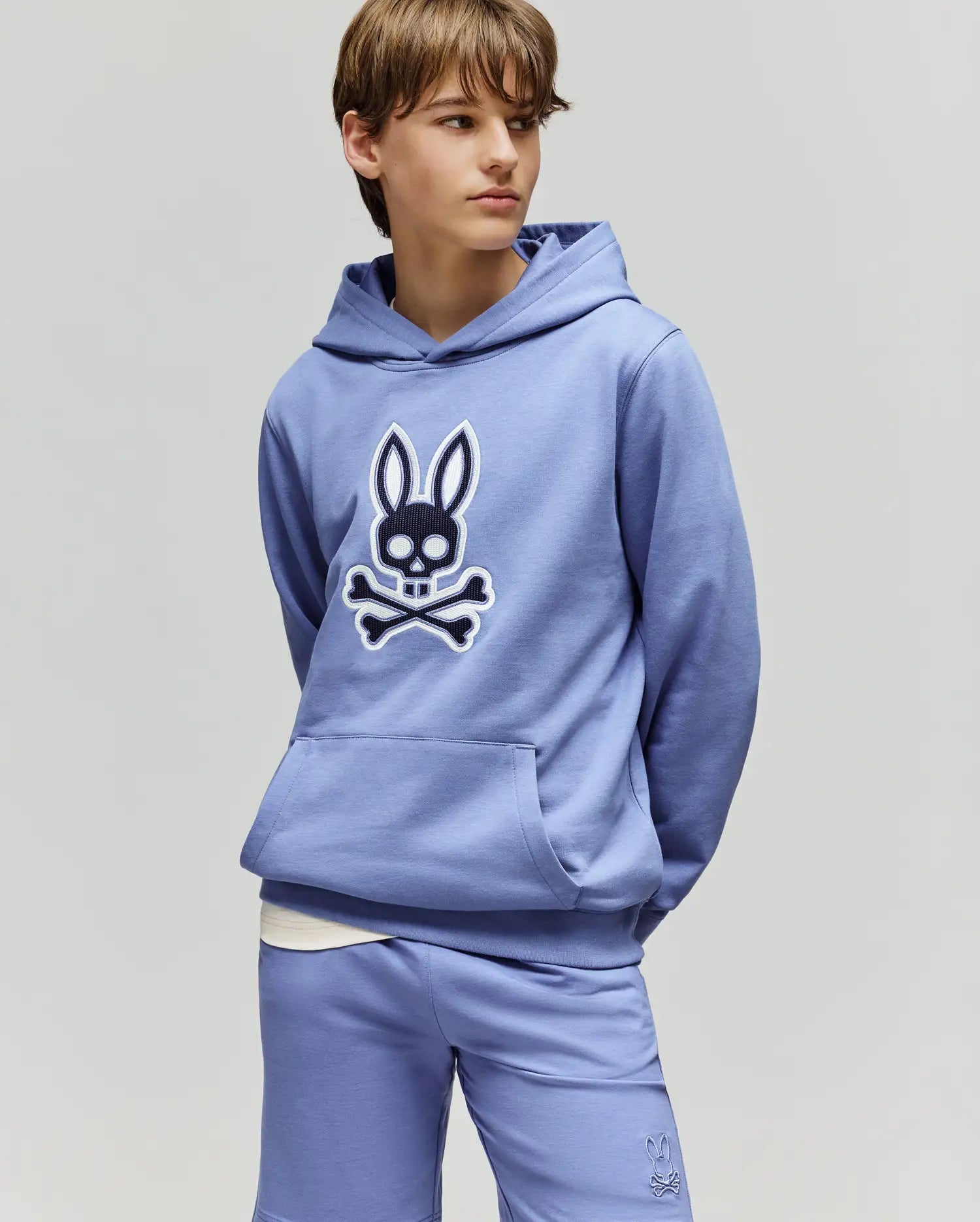 A teenage boy wears a light purple Psycho Bunny KIDS RYDER EMBROIDERED BUNNY PATCH HOODIE (KT0500018) and matching shorts, hands in pockets, gazing to the side against a plain background.