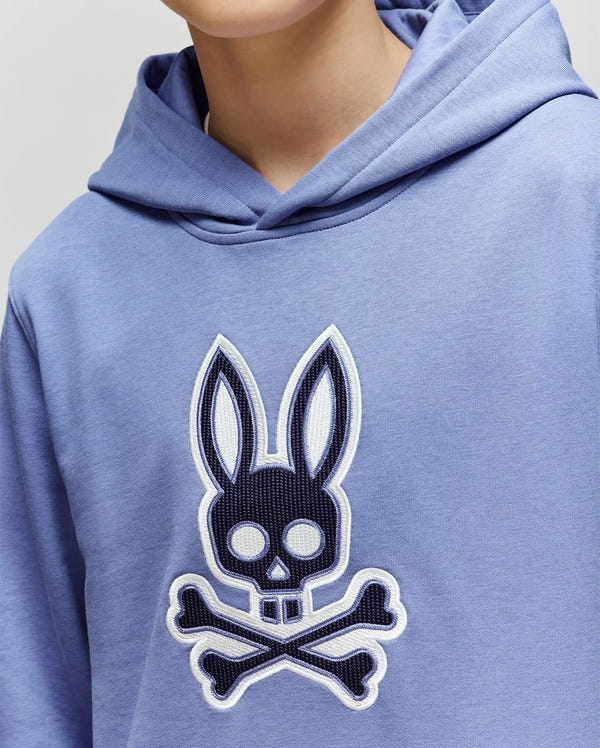 A child sports the Psycho Bunny KIDS RYDER EMBROIDERED BUNNY PATCH HOODIE (KT0500018) in light blue, featuring a large, embroidered stylized skull and crossbones Bunny patch—ideal for relaxed loungewear style.