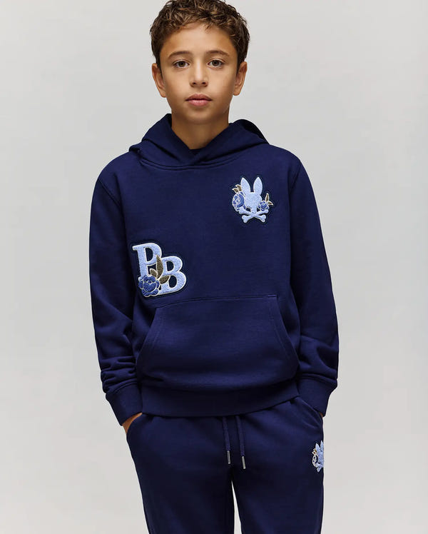 A young boy wears the Psycho Bunny KIDS AUGUSTE HOODIE (KT0500019), a navy blue regular fit cotton fleece hoodie with matching sweatpants, both featuring cartoon-style patches like a rabbit and "PB," standing with hands in his pockets.