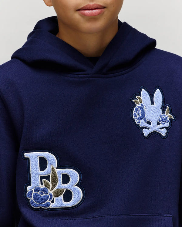 A child wears the Psycho Bunny KIDS AUGUSTE HOODIE (KT0500019), a navy blue cotton fleece hoodie featuring large "PB" and chenille floral patches on the chest, plus a bunny head with crossed bones and flowers on the upper left.