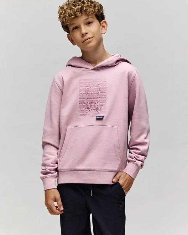 A boy with curly hair wears a Psycho Bunny KIDS GRANT HOODIE (KT0500020) in light pink, made from a premium cotton blend with an embossed bunny skull and crossbones on the chest, paired with dark pants against a light gray background.