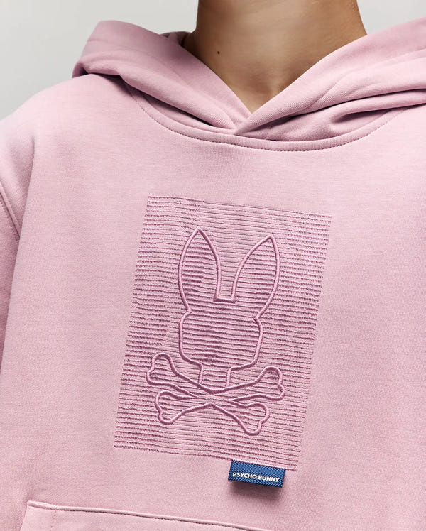 A child wears the Psycho Bunny KIDS GRANT HOODIE (KT0500020) in light pink, crafted from a premium cotton blend, with an embossed bunny and crossbones logo on the chest and a small navy "Psycho Bunny" tag near the kangaroo pocket.