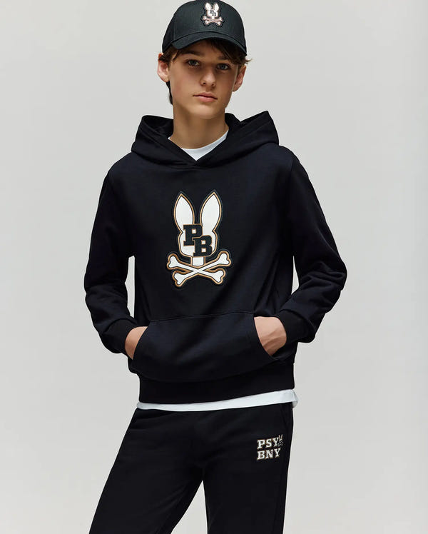 A boy wears the Psycho Bunny KIDS WADE CORD PATCH HOODIE (KT0500031) and sweatpants, both with matching PB bunny and crossbones logos, plus a black cap with an embroidered Bunny patch. Made from a premium cotton blend for comfort.
