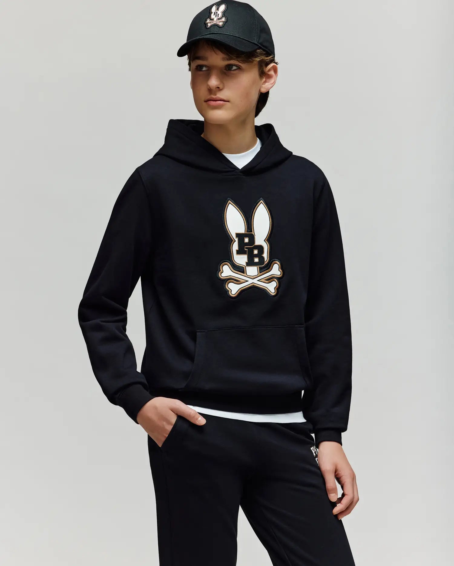 A teen boy wears the Psycho Bunny KIDS WADE CORD PATCH HOODIE (KT0500031), showcasing its premium cotton-blend fabric and signature Bunny patch with crossbones, paired with black pants and a black baseball cap.