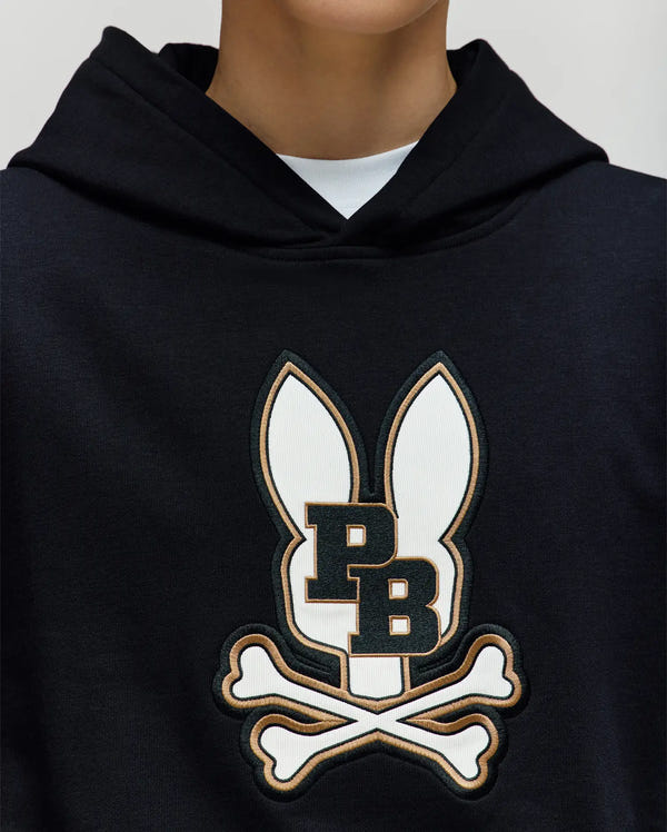 A child wears the Psycho Bunny KIDS WADE CORD PATCH HOODIE (KT0500031)—a black cotton-blend hoodie with an embroidered Bunny patch, large ears, "PB" lettering, and crossed bones—layered over a white shirt. The face is not visible.
